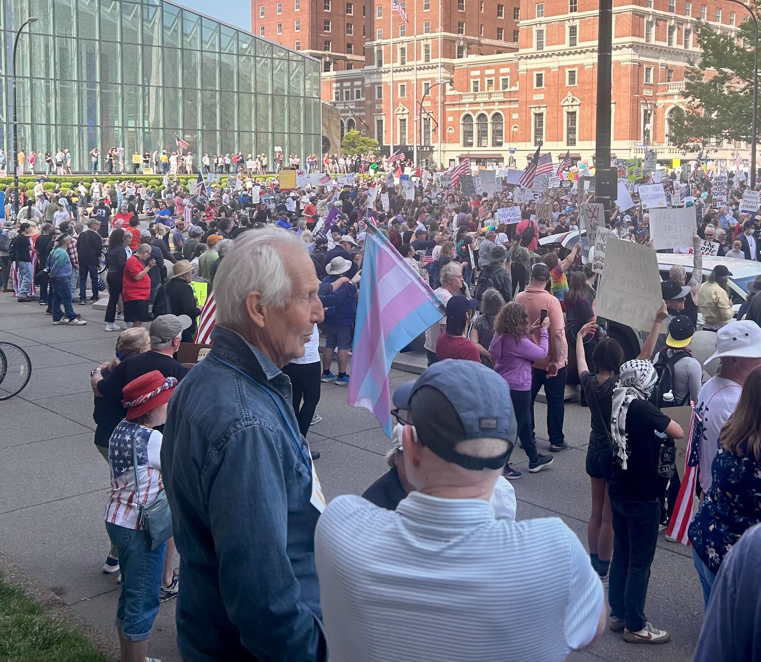 A large, diverse crowd gathers at a No Kings Day rally in Buffalo, NY, holding protest signs and American flags. A visible transgender pride flag waves in the foreground, symbolizing inclusion and solidarity. The scene, set against historic buildings and a modern glass structure, reflects the communal energy and democratic spirit described in Amber Chandler’s blog “No Kings Day: This Is What Democracy Looks Like.”